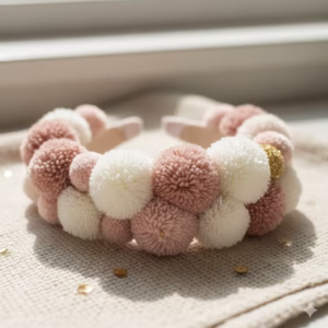 A handcrafted Pom Pom Headband featuring soft pink, creamy white, and subtle gold glitter pom poms, artfully arranged on a light-coloured woven fabric, bathed in bright, natural light. This lovely example of Pom Pom crafts showcases the charm of diy Pom Poms.